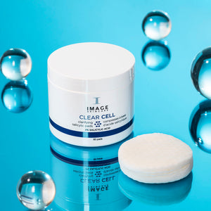 CLEAR CELL salicylic clarifying pads | IMAGE Skincare jar with white pads displayed on a reflective blue surface surrounded by translucent spheres, emphasizing skincare and cleansing.