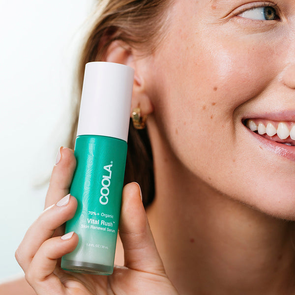 Woman holding a green bottle of Vital Rush™ Skin Renewal Serum | COOLA near her smiling face, showcasing clear, glowing skin in a bright setting.