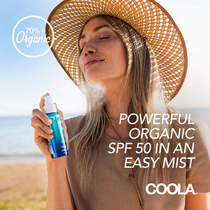 Woman wearing a wide-brimmed hat spraying Classic Face Sunscreen Mist SPF 50 | COOLA on her face outdoors in a sunny beach setting, highlighting organic skincare and sun protection.