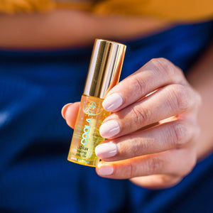 Hand holding a small transparent bottle of Classic Liplux® Organic Hydrating Lip Oil Sunscreen SPF 30 with a gold cap, against a blurred blue background.