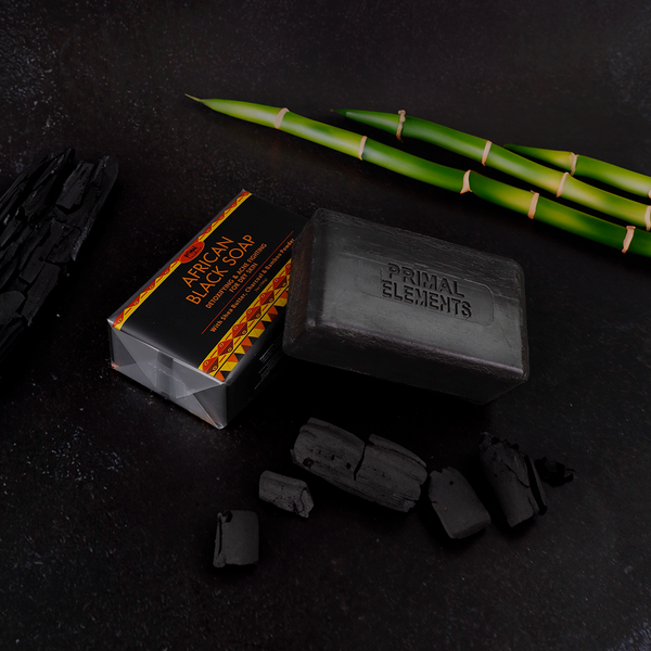 Luxury African Black Soap | Primal Elements bar with natural charcoal pieces and green bamboo stalks on a dark surface, highlighting skincare and wellness benefits.