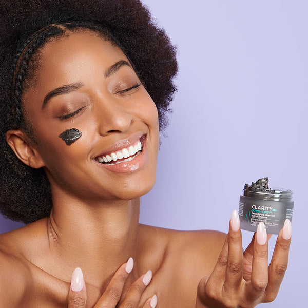 Smiling woman with glowing skin applies Down + Dirty™ Detoxifying Charcoal MicroExfoliant | ClarityRx on her cheek, holding an open jar of the black exfoliant against a light purple background.