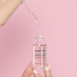 Hand holding a clear glass bottle of Glimmer Of Hope™ Shimmering Facial Oil | ClarityRx with a dropper releasing a droplet above the bottle against a soft pink background.