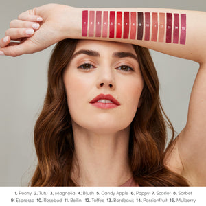 A woman with natural makeup holds her arm above her head, displaying 15 numbered swatches of ColorLuxe Hydrating Cream Lipstick | Jane Iredale in various shades from pinks to deep berries, showcasing the color range.