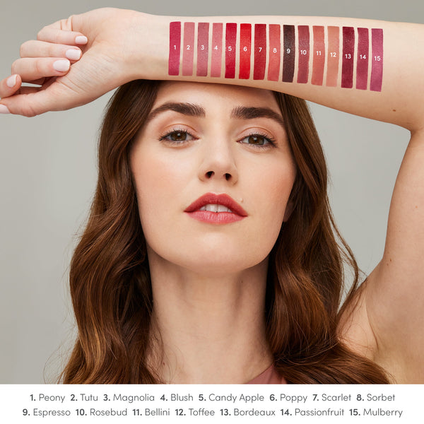 A woman with natural makeup holds her arm above her head, displaying 15 numbered swatches of ColorLuxe Hydrating Cream Lipstick | Jane Iredale in various shades from pinks to deep berries, showcasing the color range.