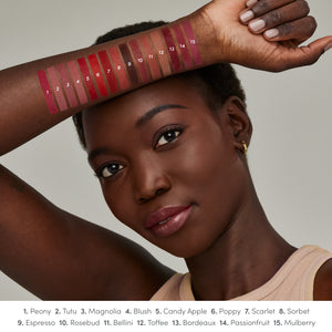 A woman with deep brown skin tone shows arm swatches of 15 rich, creamy lipstick shades from ColorLuxe Hydrating Cream Lipstick | Jane Iredale, ranging from pinks to reds and browns.