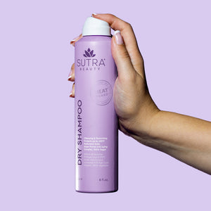 Hand holding a lavender aerosol can of Heat Guard Dry Shampoo | SUTRA against a light purple background, emphasizing the product's clean and modern design.