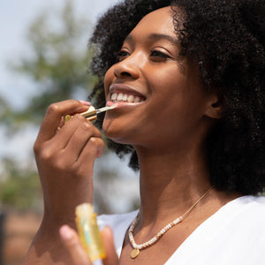 Woman outdoors smiling while applying Classic Liplux® Organic Hydrating Lip Oil Sunscreen SPF 30, showcasing skincare and sun protection in a natural setting.