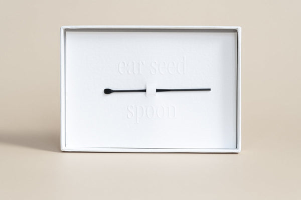 Minimalist white packaging displaying a black spoon tool accessory for ear seeds, part of The Foundation - 24K Gold Ear Seed | Vie Healing wellness product.