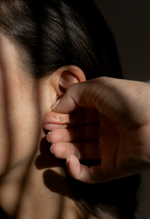 Close-up of a person gently touching their ear adorned with The Foundation - 24K Gold Ear Seed | Vie Healing, showcasing its small, elegant gold design for wellness and skincare.