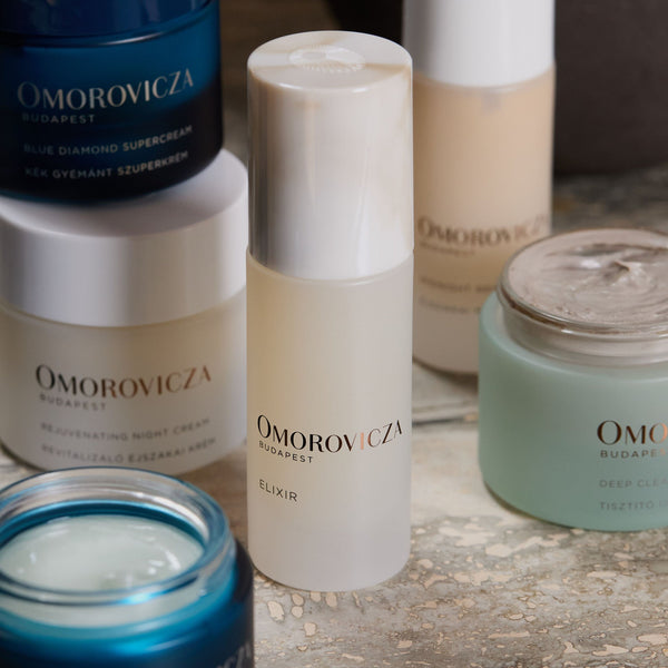 Elixir | Omorovicza skincare bottle surrounded by various luxury skincare products on a textured surface promoting wellness and body care.
