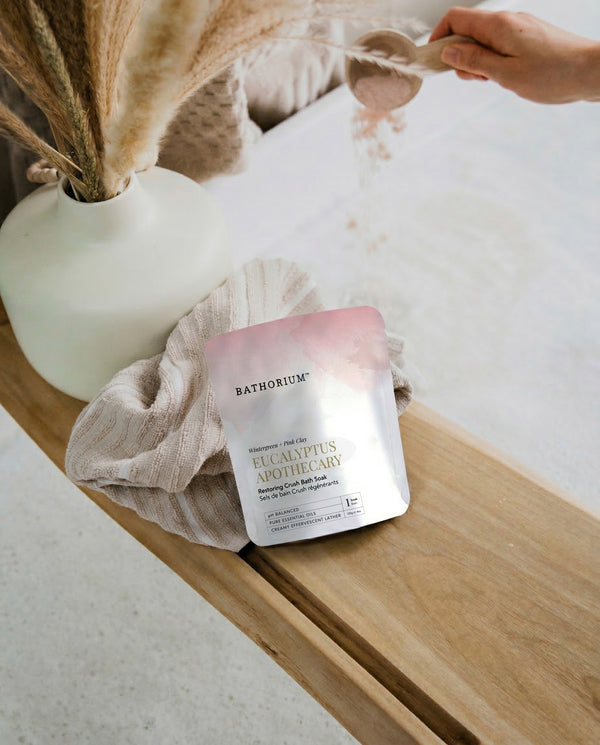 Eucalyptus Apothecary Crush | Bathorium pouch resting on a folded towel beside a white vase while a spoon pours bath powder for a calming wellness ritual