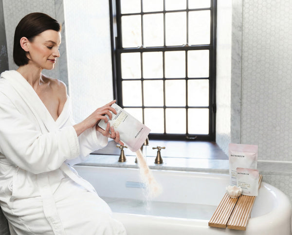 Woman in a white robe pours bath soak into a tub beside Eucalyptus Apothecary Crush | Bathorium packaging and a bamboo bath caddy for relaxing wellness