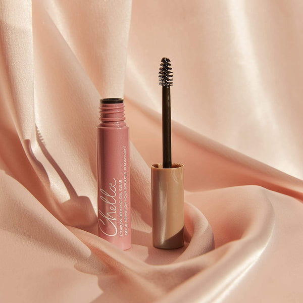 Open tube of Eyebrow Defining Gel Clear | Chella with a spoolie brush resting on soft pink satin fabric background, highlighting clear eyebrow gel product for grooming and styling.