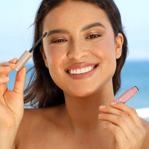 Smiling woman applying eyebrow gel with a spoolie brush near a beach, showcasing natural beauty and grooming using Eyebrow Defining Gel Clear | Chella.