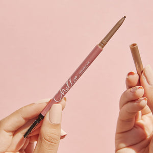 Hand holding an eyebrow pencil with spoolie brush and cap removed on a soft pink background, featuring Eyebrow Pencil | Chella in a natural brown shade.
