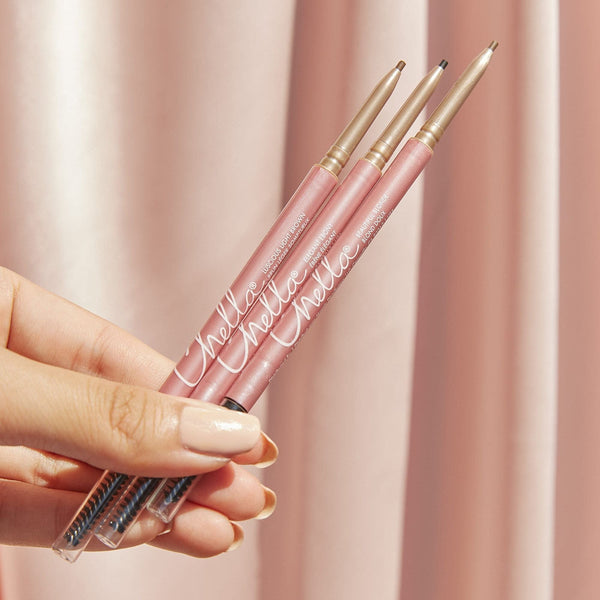 Hand holding three pink Eyebrow Pencil | Chella products with gold accents against a soft pink background, highlighting precision tips and spoolie brushes for eyebrow grooming.