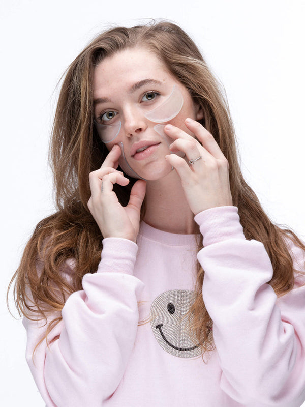 Young woman wearing clear SiliconeFusion™ Eyes & Mouth Patches | Dermaclara under her eyes, demonstrating skincare wellness and wrinkle reduction with a soft pink sweatshirt.