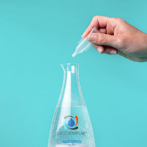 Hand holding an Activator Capsules 25ct box 12oz/3mL | Force of Nature capsule dropping liquid into a clear multi-purpose cleaner bottle against a turquoise background.