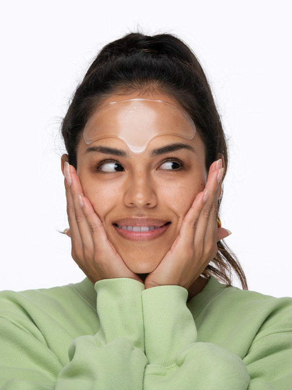 Young woman wearing a clear SiliconeFusion™ Forehead Patch | Dermaclara on her forehead, gently touching her face and smiling, promoting skincare and wellness.