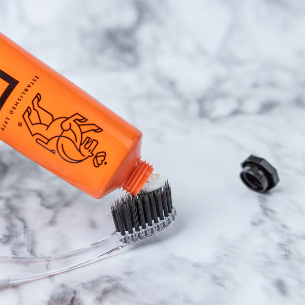 An orange tube of Linämel Toothpaste | LINHART dispensing white toothpaste onto a toothbrush with black bristles on a marble surface.
