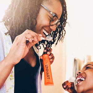 Smiling woman holding a tube of Linämel Toothpaste | LINHART and feeding a child a small amount of toothpaste on a toothbrush in a bright, cheerful setting.