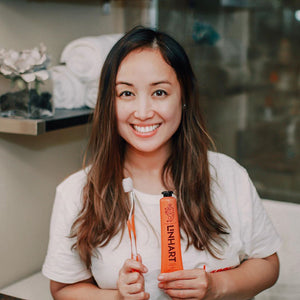 Smiling woman holding a toothbrush and a tube of Linämel Toothpaste | LINHART in a bright bathroom setting, promoting oral care and wellness.