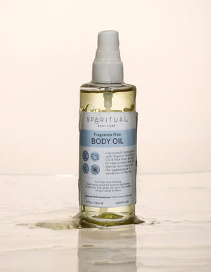 Clear plastic pump bottle of Fragrance Free Body Oil | Sparitual with light yellow oil and blue label standing on wet surface with spilled oil around it, promoting skincare and body care.