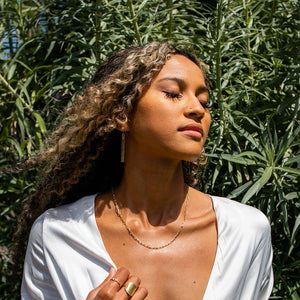 Woman with curly hair wearing a Frolic Paperclip Necklace /silver tone | Purpose Jewelry outdoors, eyes closed, dressed in white with greenery background.