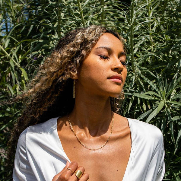 Woman with curly hair wearing a Frolic Paperclip Necklace /silver tone | Purpose Jewelry outdoors, eyes closed, dressed in white with greenery background.
