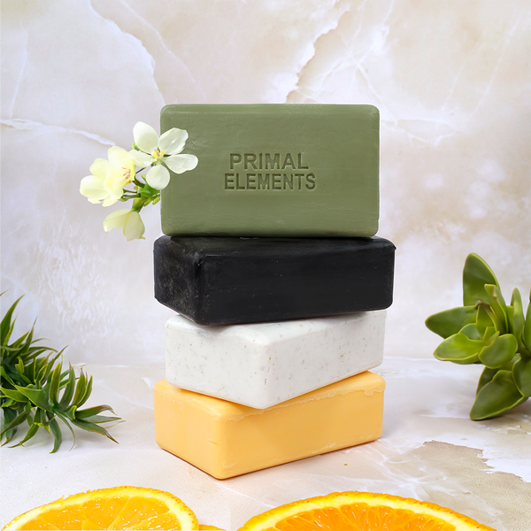 Four artisan soap bars stacked with the top green bar embossed with Primal Elements, surrounded by fresh flowers, greenery, and sliced oranges showcasing Luxury African Black Soap | Primal Elements for skincare and body care.