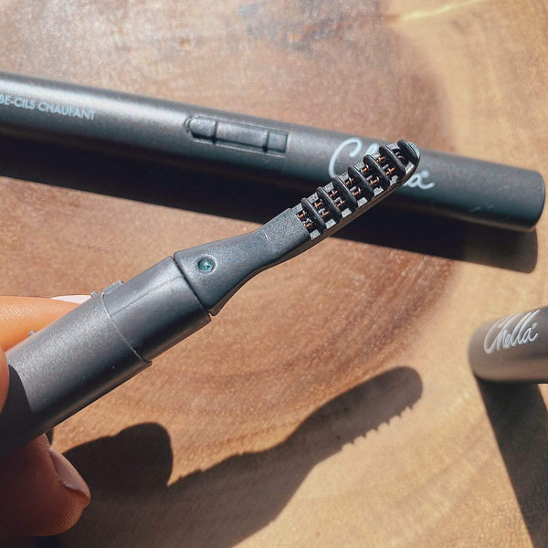 Heated Eyelash Curler | Chella held close highlighting the heated comb teeth with the product body and cap resting on a wooden surface in natural light.