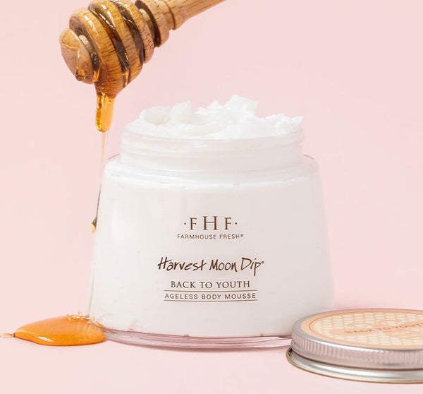 Open jar of white creamy body mousse labeled Harvest Moon Dip® | FarmhouseFresh with honey dripping from a wooden honey dipper above it on a soft pink background.