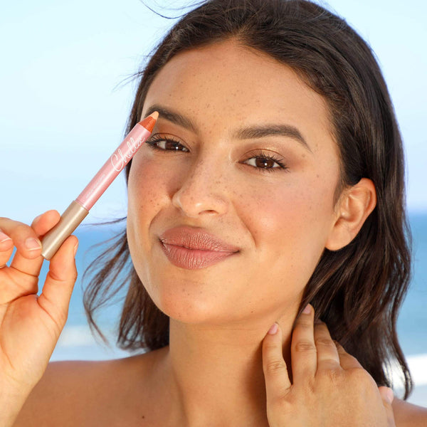 Woman with natural makeup holding a peach-toned Highlighter Pencil | Chella near her eye, showcasing a radiant and glowing skincare look against a soft blue background.