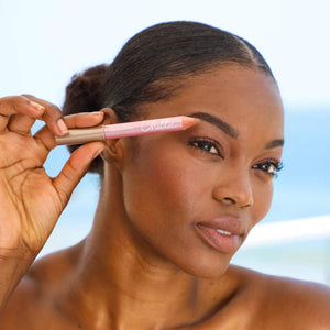 A woman with smooth skin holding a pink Highlighter Pencil | Chella near her eyebrow, demonstrating natural skincare and beauty enhancement by the seaside.