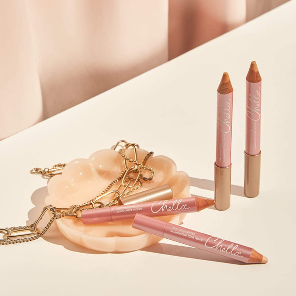 Three Highlighter Pencil | Chella products in soft pink packaging displayed on a light surface with a decorative dish and gold chain, styled for skincare and wellness aesthetic.