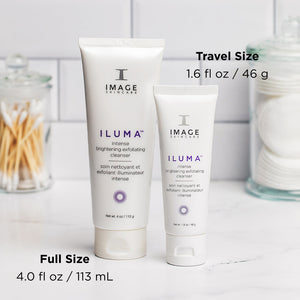 Two white tubes of ILUMA intense brightening exfoliating cleanser | IMAGE Skincare displayed on a countertop with one full size and one travel size, alongside cotton pads and cotton swabs in glass containers.