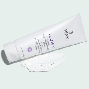 Tube of ILUMA intense brightening exfoliating cleanser | IMAGE Skincare lying next to a smear of the white exfoliating cleanser with visible exfoliating particles on a light background.