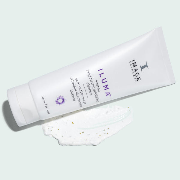 Tube of ILUMA intense brightening exfoliating cleanser | IMAGE Skincare lying next to a smear of the white exfoliating cleanser with visible exfoliating particles on a light background.