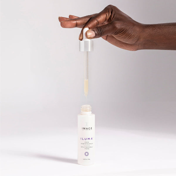 A dark-skinned hand holds a dropper releasing serum above an open bottle of ILUMA® intense brightening serum | IMAGE Skincare on a plain background.