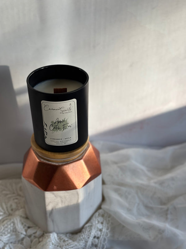 Black container candle with wood wick labeled Soft Crackling Wood Wick Candle | CommonScents, placed on geometric copper and white stand with soft natural lighting.