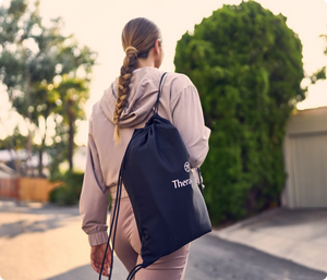 Woman walking outside wearing a light jacket and carrying a black drawstring bag with Therabody branding, promoting wellness and recovery with JetBoots Prime | Therabody.