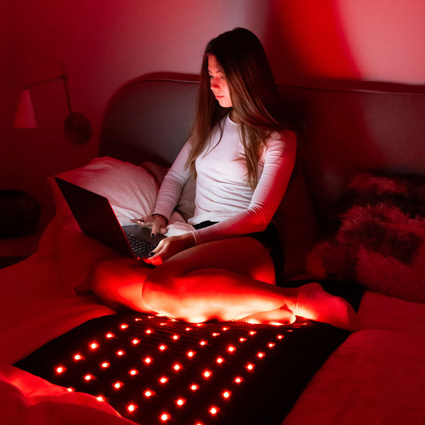 Woman sitting on bed using laptop with red light therapy from Jet Series - PEMF Far Infrared Heating Mats | HealthyLine for wellness and recovery care at home
