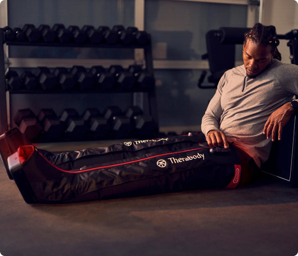 A man using JetBoots PRO Plus | Therabody compression recovery device on his legs in a gym setting with dumbbells in the background, promoting wellness and recovery.