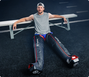 Man sitting on the ground with legs extended while wearing JetBoots PRO Plus | Therabody recovery devices on both legs in a sports setting.