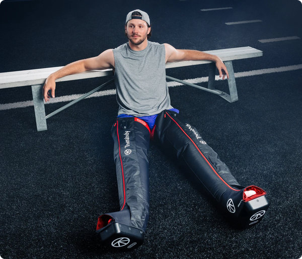 Man sitting on the ground with legs extended while wearing JetBoots PRO Plus | Therabody recovery devices on both legs in a sports setting.