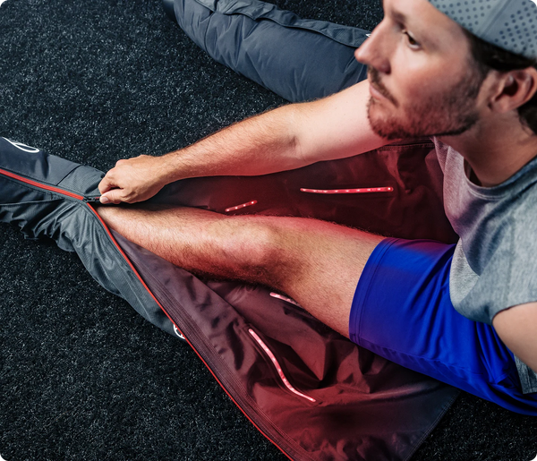 Man sitting on a black surface while using JetBoots PRO Plus | Therabody compression recovery boots with red LED lights activated on his leg.