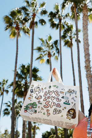 White tote bag with colorful kawaii-style spa-themed illustrations and the text Live Love Spa Kawaii Event Bag | Live Love Spa held up outdoors against a backdrop of tall palm trees and clear blue sky.