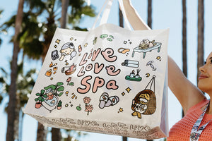 A person holds a white Live Love Spa Kawaii Event Bag | Live Love Spa featuring cute spa-themed illustrations and pastel colors outdoors with palm trees in the background.