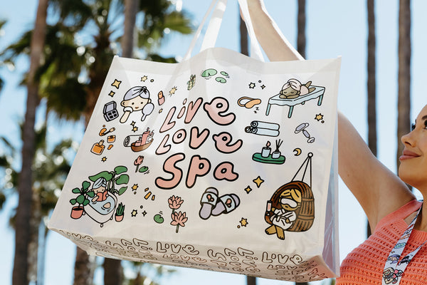 A person holds a white Live Love Spa Kawaii Event Bag | Live Love Spa featuring cute spa-themed illustrations and pastel colors outdoors with palm trees in the background.